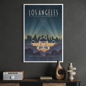 A framed Los Angeles cityscape poster hangs on a dark gray wall above a console table with decorative objects including a radio, books, and vases. The poster features stylized artwork of the Los Angeles skyline and Griffith Park under a starry night sky, with the text 'LOS ANGELES' at the top.