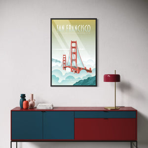 A framed vintage-style travel poster of San Francisco featuring the Golden Gate Bridge, displayed on a light gray wall above a red and blue sideboard with decorative objects and a red lamp.