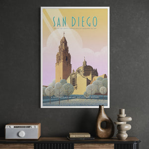 A framed vintage-style travel poster of San Diego featuring the Cathedral bell tower and surrounding architecture, hung on a dark gray wall above a console table with decorative objects.