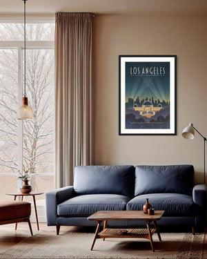 Framed Los Angeles Griffith Observatory travel poster hanging above a modern blue sofa with wooden coffee table and side tables in a cozy living room with large window showing snowy trees outside.