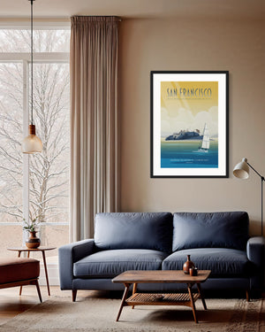 Framed San Francisco Alcatraz poster displayed above a dark blue sofa in a warm, softly lit living room with large windows.