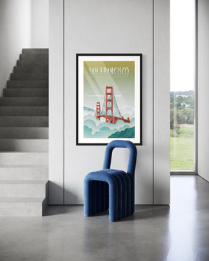 Framed San Francisco Golden Gate Bridge poster hanging on a light gray wall beside modern concrete stairs, with a sculptural blue velvet accent chair in the foreground. Minimalist, contemporary interior décor setting.